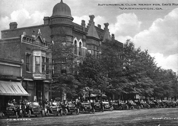 View across road toward the Automobile Club, arranged in a long line of automobiles along a tree-lined commercial street. Caption reads: "Automobile Club Ready for Day's Ride, Washington, GA."