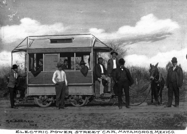 A group of men pose near an electric power street car first used in 1832. A mule is hitched to the streetcar. Caption reads: "Electric Power Street Car, Metamoros, Mexico."