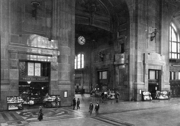 Slightly elevated view of people walking through the main lobby of the Union Railroad Station, built in 1914. A fruit and candy shop with a soda fountain is on the left, along with a six-foot wide clock hanging in the Station's central arch.