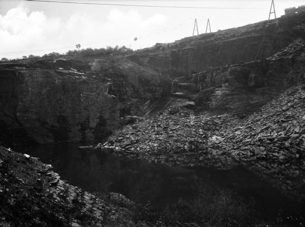 The slate industry was begun in Bangor by Robert M. Jones in 1848. The region became a major world center for slate. The view is from near the bottom of the quarry, which is filled with water, looking up a steep side of the quarry with industrial equipment and buildings near the top. Pulley blocks are suspended from cables attached to towers.