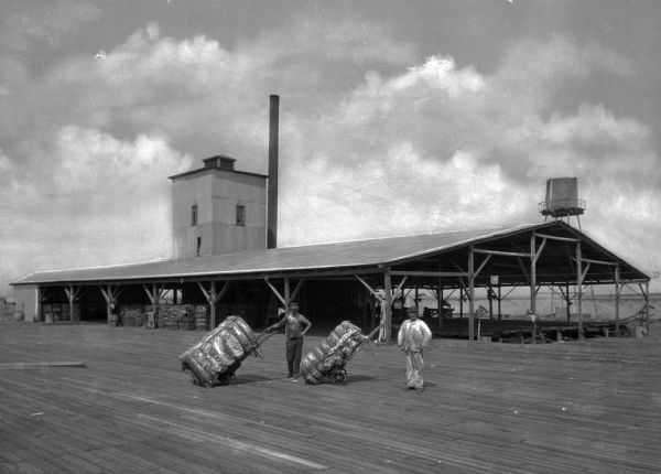Two men hold carts of cotton bales in front of the warehouse of a cotton compress.