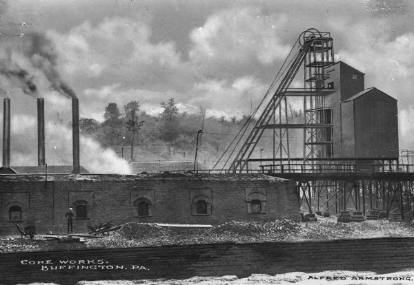 The roofs and smokestacks of the power house and hoist house of Buffington Coke Works are behind the coke ovens which are in the foreground. Buffington Coke Works contained 426 bee-hive coke ovens by 1910. Caption reads: "Coke Works, Buffington, PA."