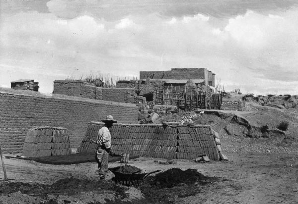 A man works in an adobe brickyard with adobe structures in the background.