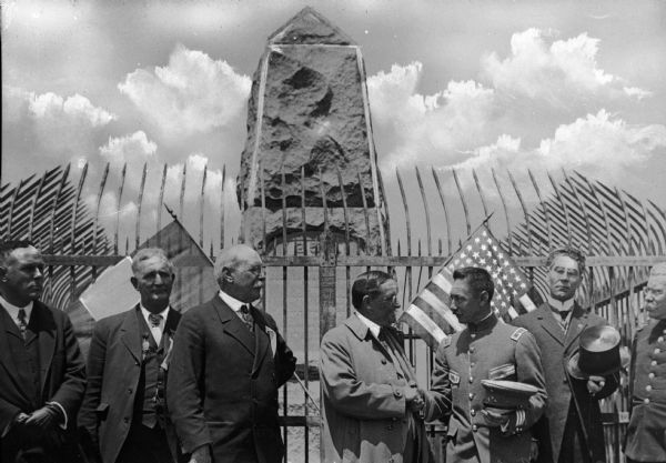 Officials greet one another in front of Boundary Monument 258 between the United States and Mexico. A protective fence is behind the men.