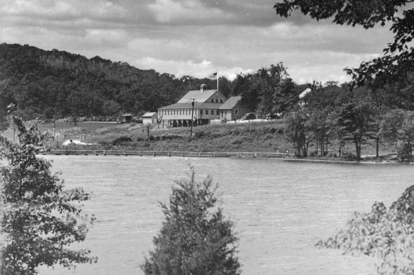 A view of a Northland Camp building from across Lake Iliff. The camp was operated as an auxiliary of the German-American Bund, the American Nazi Organization. The United States flag waves on a pole behind the building, and a hill rises in the background.