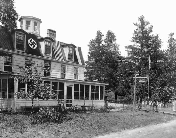 A front view of a house with a swastika, flag, and symbol of the Nazis of Germany at Camp Sigfried. In front of the house, a flag waves on a pole and a sign reads, "Camp Sigfried."