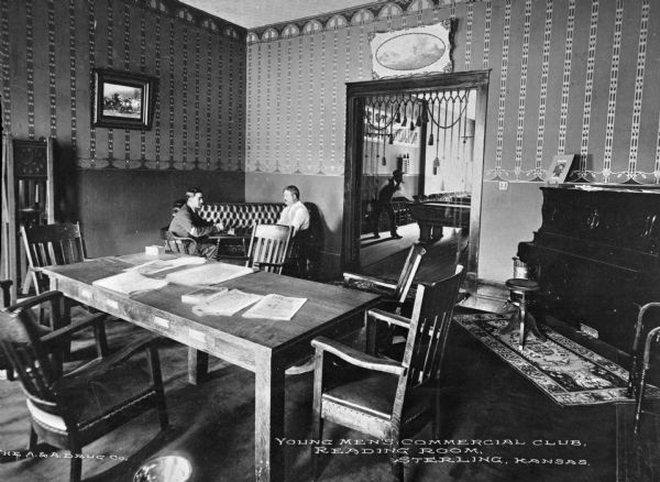 A view of two men sitting in the back of the room, and books and papers on top of a table, taken from across the reading room of the Young Men's Commercial Club. A piano is in the right corner, and paintings line the walls. A man plays pool in a room in the background while smoking a cigar. Caption reads: "Young Men's Commercial Club, Reading Room, Sterling, Kansas."