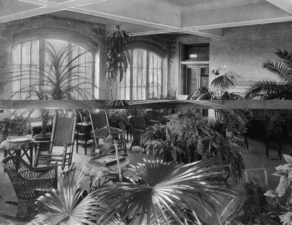 A view of chairs, tables, and plants in the Grand Lodge Hall Conservatory of the Masonic Temple. A fireplace and windows are in the background.