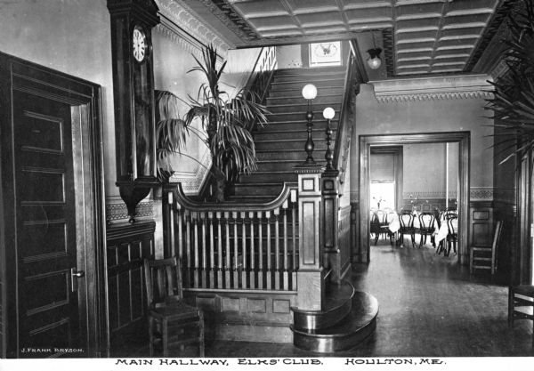 A view of the main hallway of the Elks Club, founded in 1868, showing the entrance to the dining room and the stairway upstairs. A clock hangs on the wall near a door, and plants decorate the space. Caption reads: "Main Hallway, Elks' Club, Houlton, ME."