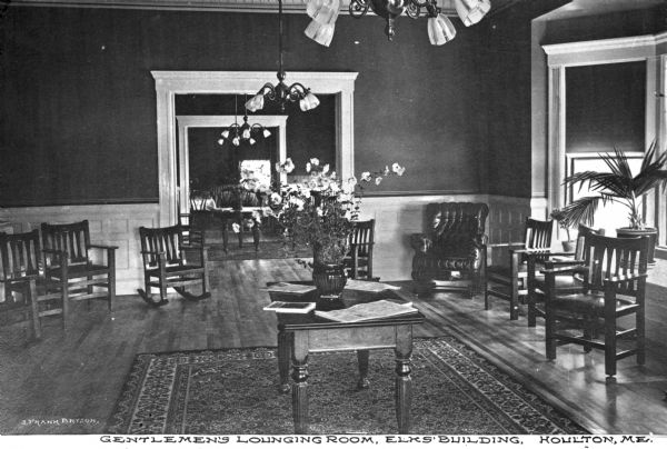 A view of chairs and a table with flowers and papers in the gentlemen's lounging room of the Elks Club, founded in 1868. Caption reads: "Gentlemen's Lounging Room, Elks' Building, Houlton, ME."