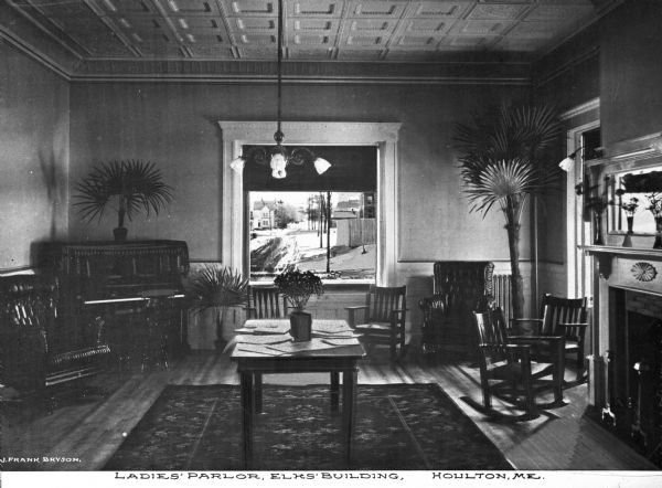 A view of a piano, chairs, a table, and a fireplace in the ladies parlor of the Elks Club, founded in 1868. An open window shows a street scene and a home. Caption reads: "Ladies' Parlor, Elks' Building, Houlton, ME."