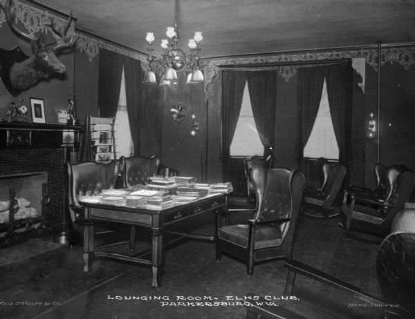 A view of a fireplace, chairs, and tables in the lounging room of the Elks Club, founded in 1868. Stacks of books lie on the table, and decorations and lighting line the walls. Caption reads: "Lounging Room, Elks Club, Parkersburg, W. VA."