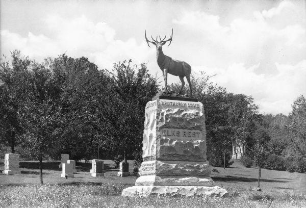 A view of an elk atop the Elks Rest Monument in Mount Washington Cemetery. Gravestones and trees can be seen in the background. The inscription on the monument reads, "Elks Rest," and "Brotherly Love."