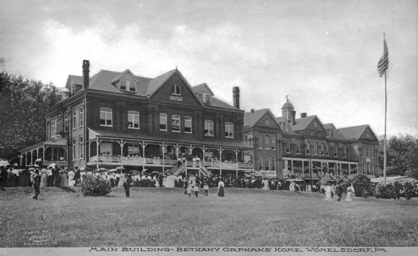A view from across the front lawn of people gathered around the main building of Bethany Orphans' Home, built in 1898. A United States flag is on a pole on the far right. Caption reads: "Main Building, Bethany Orphans' Home, Womelsdorf, PA."