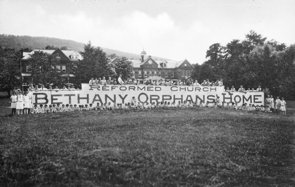 Outdoor group portrait of orphans and adults around a sign that reads, "Reformed Church Bethany Orphans' Home." The home is in the background.