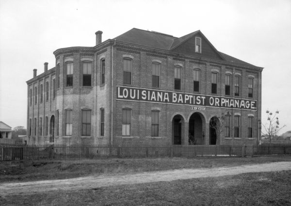 A view from across the dirt path of Louisiana Baptist Orphanage, constructed in 1903. A fence surrounds the building which has tall windows.
