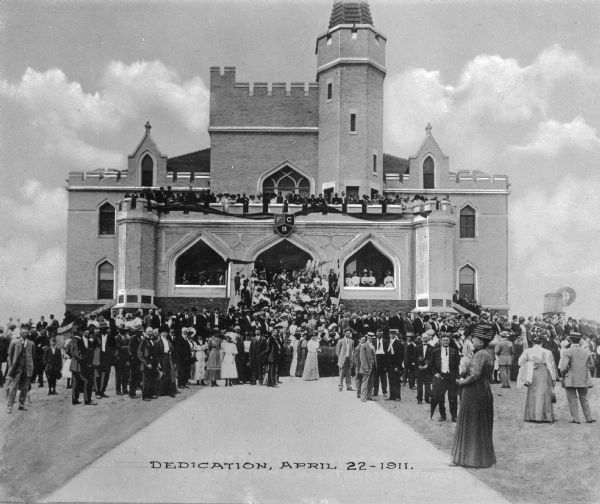 Front view of Pythian Orphans' Home and a large crowd in front of the main building. The shield over the entrance reads, "F C B," standing for the motto of the Knights of Pythias, "Friendship, Charity and Benevolence." A windmill can be seen in the background on the right. Caption reads: "Dedication, April 22 — 1911."