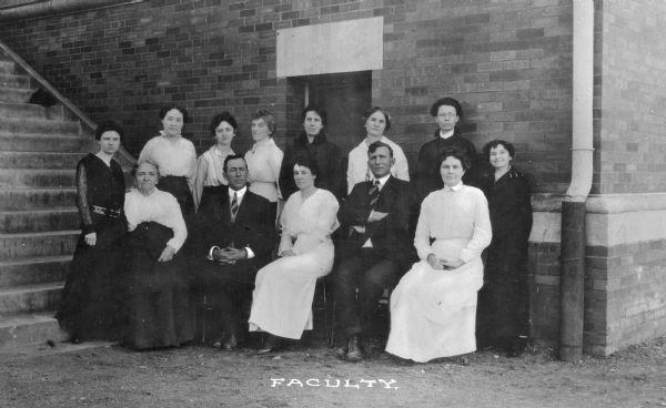 A posed group portrait of the Pythian Orphans' Home faculty. The group poses in front of a brick wall with a window, and a staircase on the left. Caption reads: "Faculty."
