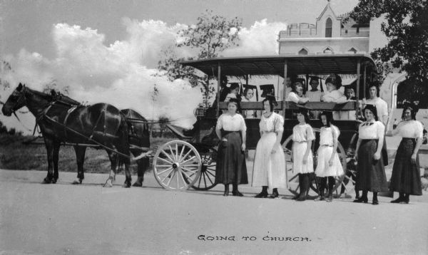 A group of women wearing tasseled commencement hats stand in front of and sit in a horse-drawn wagon, on their way to church. The Pythian Orphans' Home is in the background. Caption reads: "Going to Church."