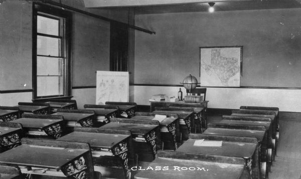 A view from the back of a classroom of desks, a globe, and a map of Texas at Pythian Orphans' Home. Caption reads: "Class Room."