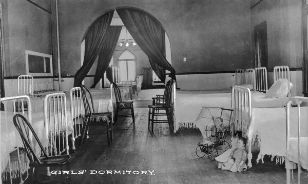 An interior view of the girls' dormitory of Pythian Orphans' Home.  Beds are lined up on both sides of the room, and draped arches lead into more rooms in the background. Dolls are on the floor at the foot of a bed on the right. Caption reads: "Girls' Dormitory."