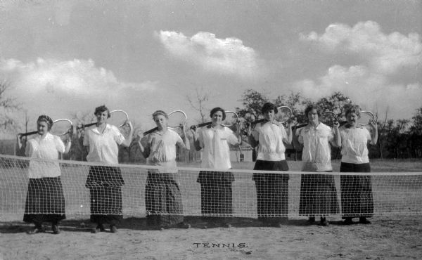 A posed portrait of women tennis players posed behind the net, holding their rackets. Pythian Orphans' Home can be seen in the background. Caption reads: "Tennis."