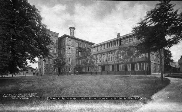 Exterior view of the Male Almshouse, a home for the aged and ill.  The brick structure is lined with windows, and two adults walk past to the left of the home. Caption reads: "Male Alms House — Blackwell's Island, N.Y."