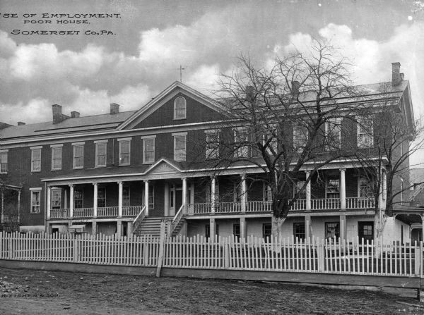 Front view of the House of Employment, or the Poor House. A fence is shown in the foreground and a staircase leads to a long porch and the front entrance of the house. Caption reads: "House of Employment, Poor House. Somerset Co., PA."