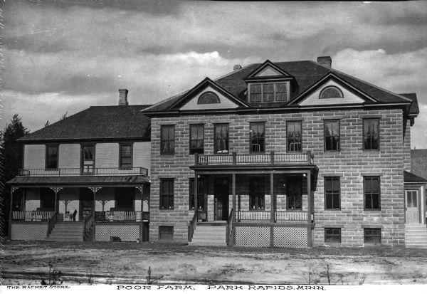 Front view of the Poor Farm. Two front porches attach to the houses with decorative columns and lattice work. One of two front doors is open, and a side door is shown on the far right. Caption reads: "Poor Farm. Park Rapids, Minn."