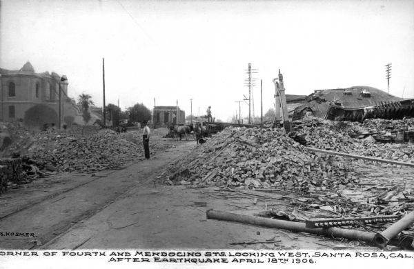 Street scene at the corner of Fourth and Medocino Streets, looking west. A man stands next to the piles of ruins, looking at the camera. A horse and carriage is in the background. Caption reads: "Corner of Fourth and Mendocino Sts. Looking West, Santa Rosa, Cal. After the earthquake of April 18th 1906."
