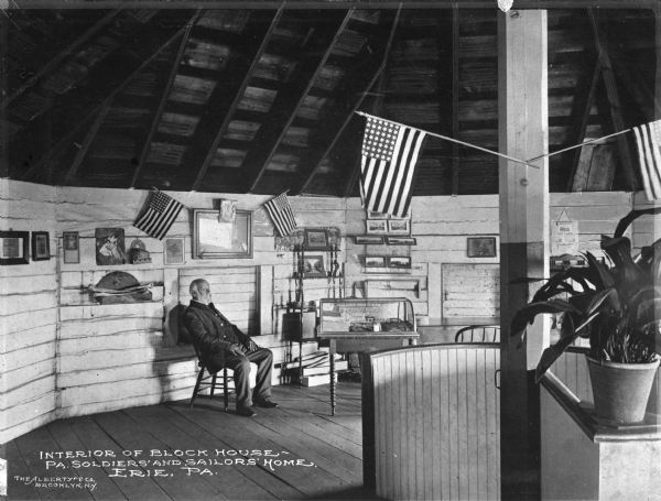 Interior view of the block house with a steep exposed ceiling. A man is siting in a chair, and United States flags adorn the columns and a framed image on the wall. There is a potted plant in the right foreground. Caption reads: "Interior of Block House — PA. Soldiers' and Sailors' Home. Erie, PA."