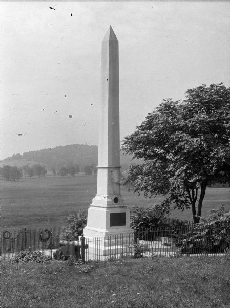 View of Waterman's Monument, a 50 foot granite obelisk erected in 1901 by the Daughters of the Revolution. The monument marks the site of the only identified grave at Valley Forge, that of Lieutenant John Waterman. A fence surrounds the memorial, and a field and trees are in the distance.