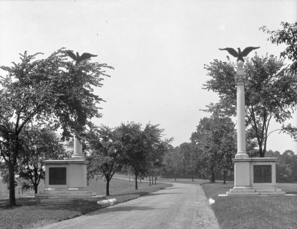 Two statues mark the entrance of the picnic area of Valley Forge National Park. The monuments consist of identical bases and an eagle on top of each column. The road curves to the left in the distance.