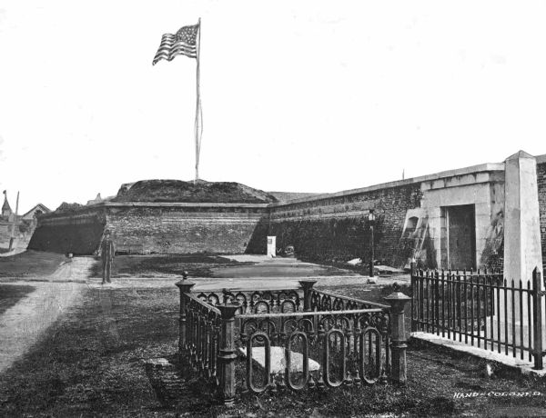 A view of a grave and a monument at Fort Sumter. A flag waves in the distance. The stone entrance can be seen on the far right.