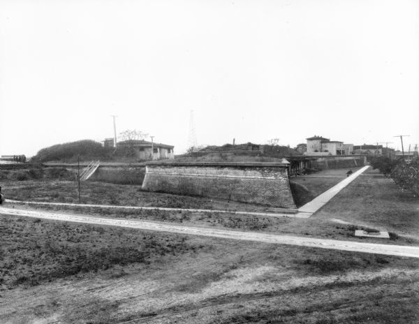 A view of the ruins of Fort Sumter from across a road showing a set of stairs leading up to a building. A path leads to other buildings which remain intact in the background on the right.