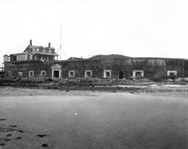 A view of Fort Moultrie from across a body of water. A doorway leads into the fort, and to the left a sign reads, "Cable Crossing Do Not Anchor." Past the wall are two dwellings.
