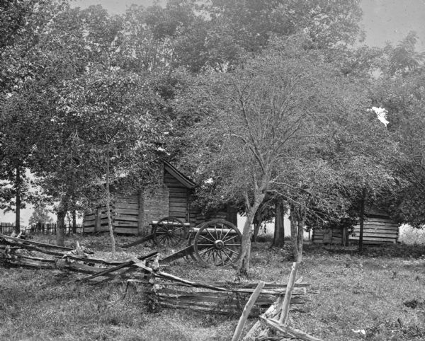 A view of cannons beneath trees behind a wooden fence. Two log cabins are in the background.