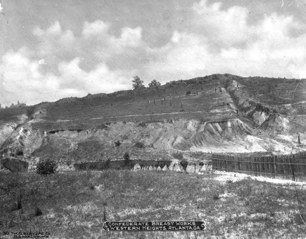 A view of Confederate breastworks, ridges in the hillsides for defense, at Western Heights. A fence divides the land in the foreground. Caption reads: "Confederate Breast Works Western Heights, Atlanta, GA."