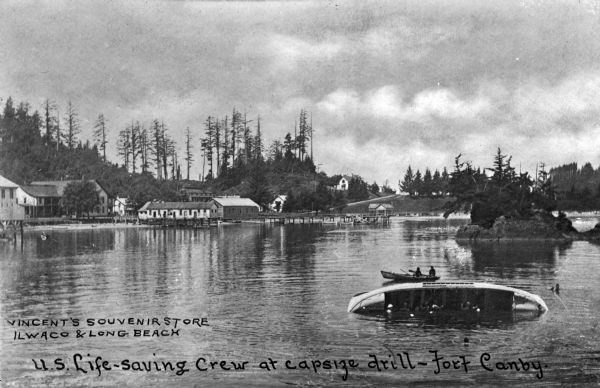 A general view of two members of the U.S. Life-saving Crew in a boat, during capsize drill. Camp facilities are on the left riverbank in a wooded area. Caption reads: "U.S. Life-saving Crew at capsize drill — Fort Canby."