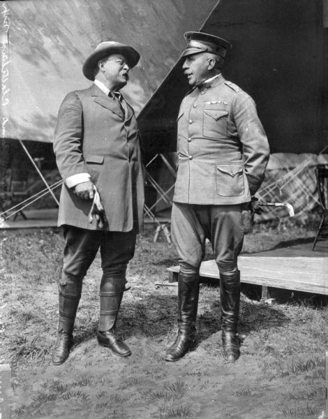 Theodore Roosevelt and General Leonard Wood conversing at a military training camp. Camp facilities are behind them.