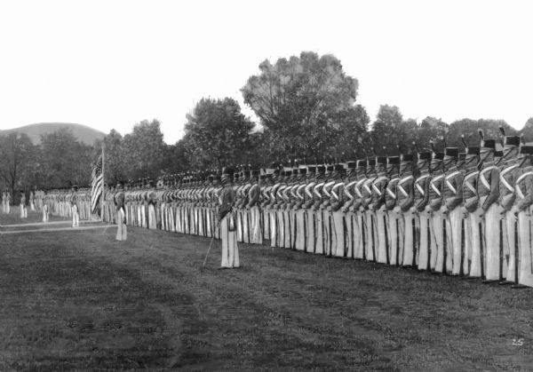 A view of soldiers in formation, wearing military uniforms at West Point U.S. Military Academy during a dress parade. A flag waves on a pole on the left.