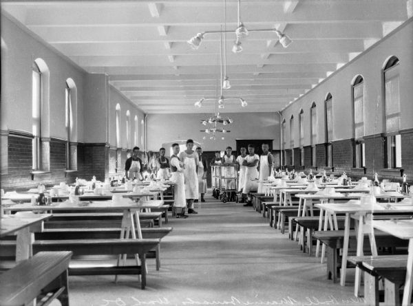 A view of the staff at the dining hall of the marine barracks.  The staff wears white aprons and prepares to serve food, and tables are set on either side of the room.