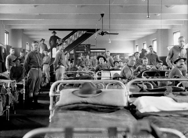 A large group of men looking at the camera, standing near their cots in a squad room of a marine barracks. A staircase leads upward on the left.