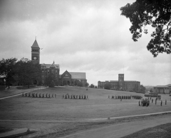 A view of a military drill on the campus of Clemson College, which opened in 1893. College buildings stand in the background, and lines of soldiers march in formation.