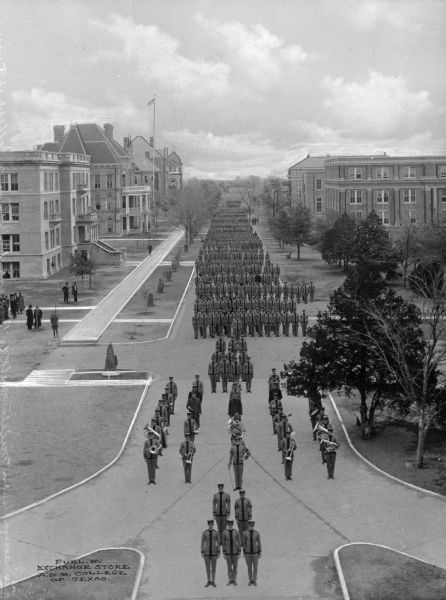 Elevated view of a Student Military Organization parade through the campus of Texas Agricultural and Mechanical College, opened on October 4, 1876. The band leads the parade. On either side are college buildings and spectators. In the background on the left a flag waves on a pole.