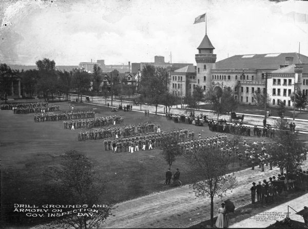 Elevated view of military formations on the drill grounds on government inspection day. A band plays for the military drill, and a flag waves on a pole on the armory. Spectators observe the drill. Caption reads: "Drill Grounds and Armory on Gov. Inspection Day."
