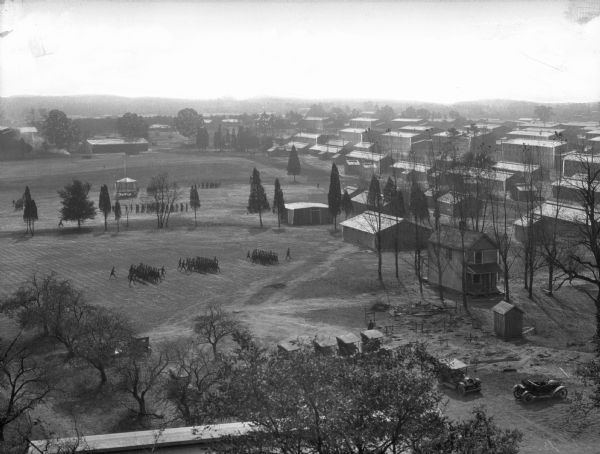 Elevated view of buildings and soldiers marching in a field. Automobiles are parked on the lower right.
