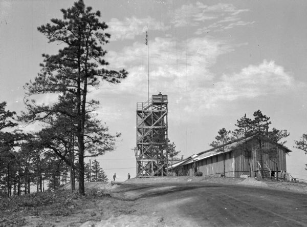 View looking uphill toward two men standing on a road near a tall lookout tower with a flag on a flagpole, and barracks at Camp Upton, active starting in 1917.