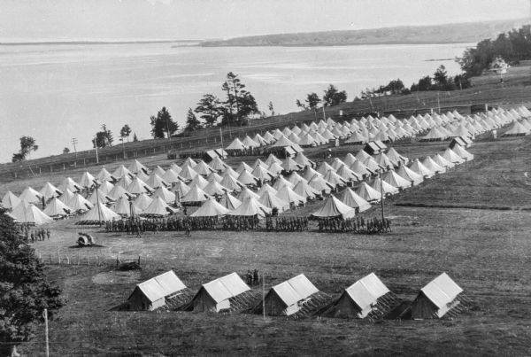Elevated view of rows of tents at a military training camp. Men in formation march in the direction of Lake Champlain.