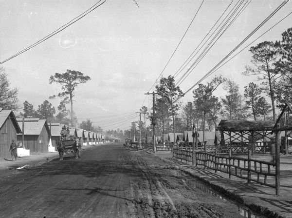 A view down Regimental Street, a dirt road at Camp Logan, shows a man in a horse-drawn vehicle, with a fence on the right, and many identical buildings on the left.